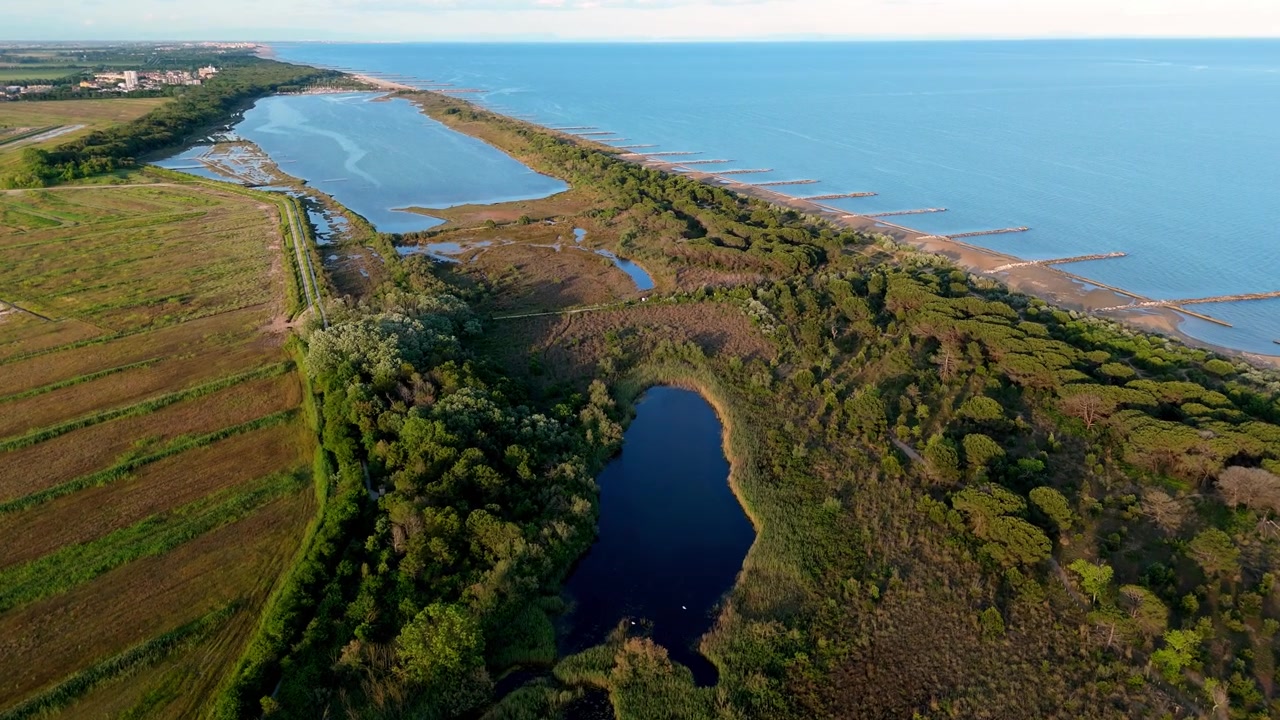 Ampia prospettiva aerea con drone dello stretto cordone litorale che separa le calme onde del mare dalle selvagge paludi lagunari e dai canali interni al tramonto, vicino a Eraclea e al fiume Piave, Italia.
