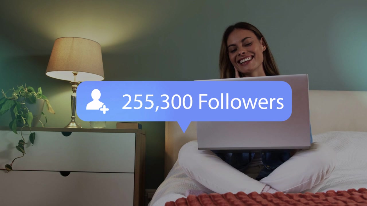 Woman sitting crosslegged on bed using laptop while social follower overlay updating causing smile
