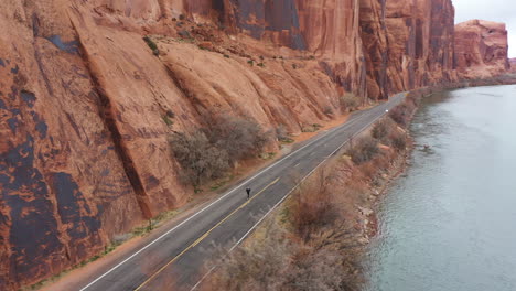 Aerial-View-of-Man-Running-Fast-of-Deserted-Road-Under-Red-Sandstone-Cliffs-of-Utah-Desert-by-Colorado-River,-Drone-Shot
