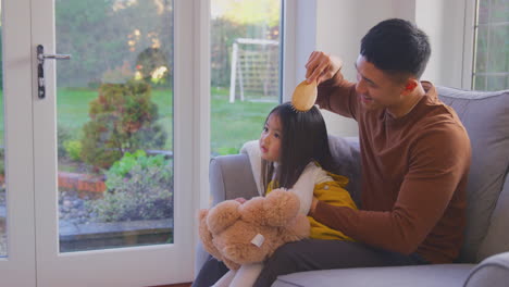 family having fun at home with dad brushing daughter's hair sitting on sofa with toy