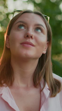 beautiful lady seated outdoors in pink top shakes head thoughtfully with a warm smile, then looks down back, sun glow from behind creates a serene, natural atmosphere