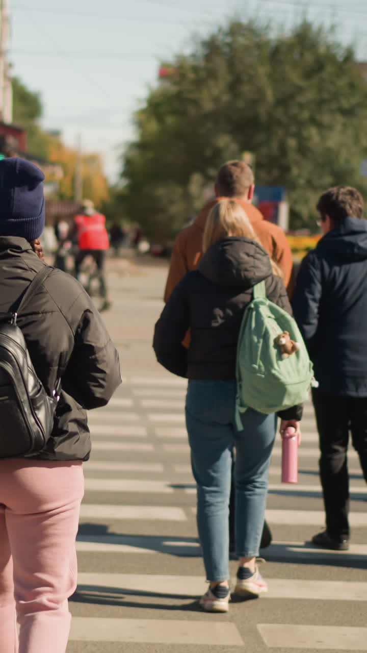 Jóvenes adultos cruzando un paso de peatones en la ciudad con mochilas. Escena callejera con mochilas y chaquetas, luz solar matutina, señal de peatones, grupo mixto de estudiantes y personas que se desplazan al trabajo, moda informal, ritmo relajado, urbano