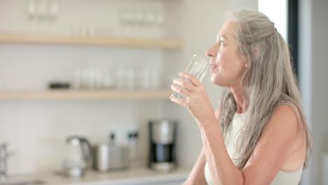 A-mature-Caucasian-woman-holding-glass-of-water,-standing-in-a-kitchen