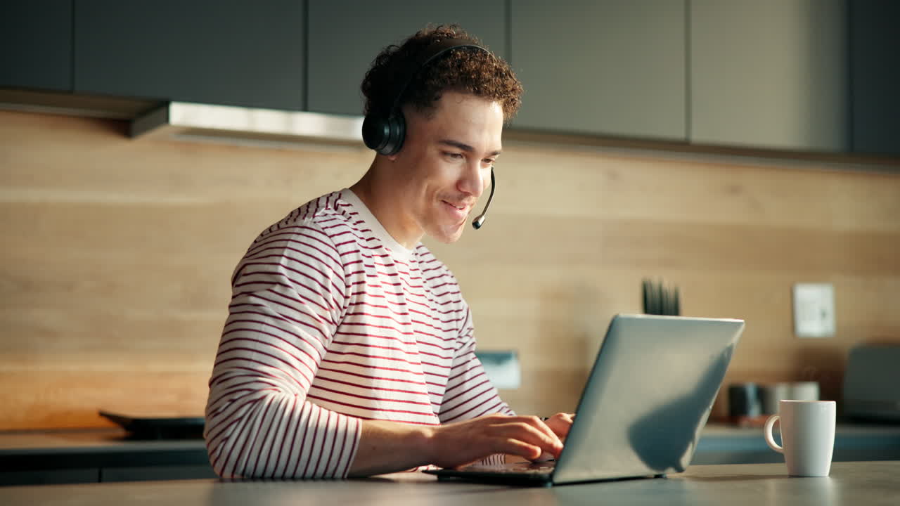 Ung mand med headset arbejder på bærbar computer i moderne køkken