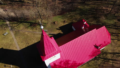 aerial view of lielvarde lutheran church at the bank of daugava river, white church with red roof, leafless trees, sunny spring day, wide birdseye shot moving forward