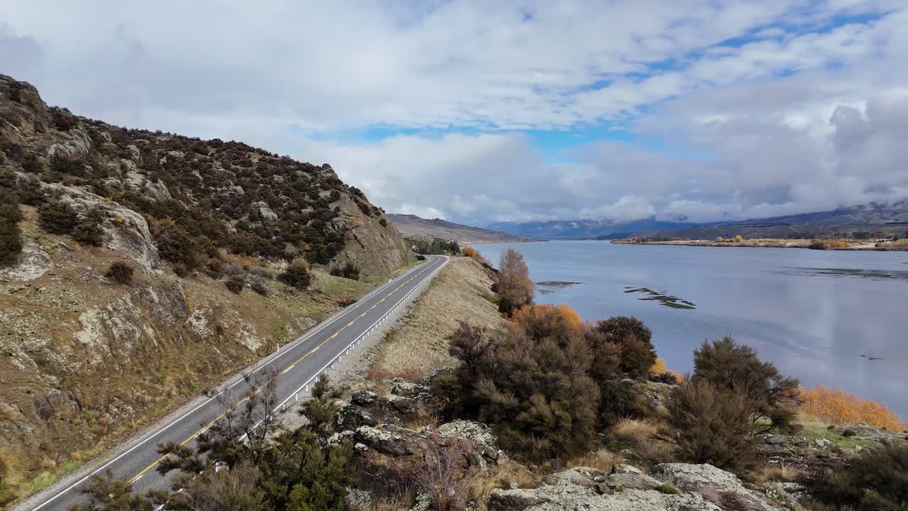 Drone glides over rocky outcrop, revealing a winding lakeside road bordered by hills, autumn trees, and reflective water under partly cloudy daylight