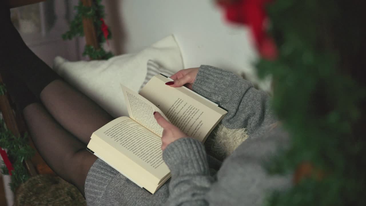 A woman wearing a gray sweater and black tights relaxes while reading a book in a cozy home setting. Warm tones and holiday decorations create a peaceful winter atmosphere