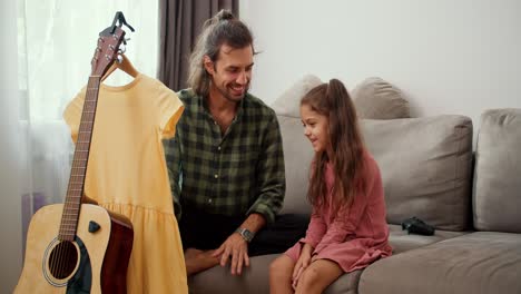 A-little-brunette-girl-in-a-pink-dress-sits-on-the-sofa-near-her-father-who-steams-a-pink-dress-using-a-special-steamer-in-a-modern-apartment.-Single-father-brunette-man-in-a-green-checkered-shirt-steams-his-daughter's-yellow-dress-at-home