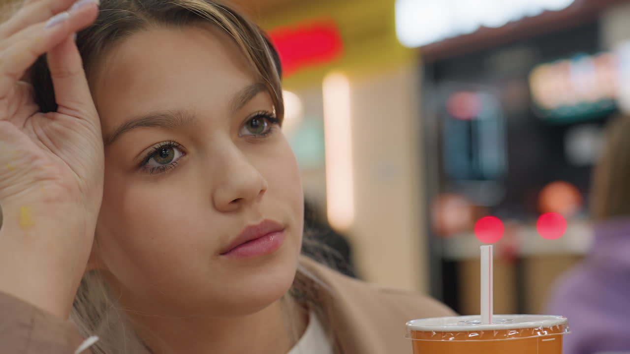 Joven influencer caucásica bebiendo una bebida de naranja en un concurrido mercado gastronómico, mirada soñadora, mano en el pelo, vaso de marca, luces bokeh cálidas, blazer elegante, maquillaje suave, ambiente de redes sociales, encuadre espontáneo.