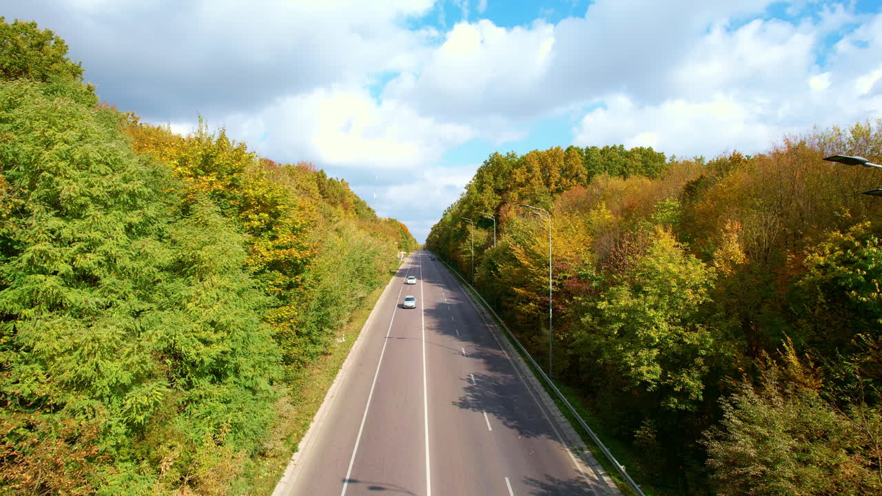 Cars riding by the highway in the woods. Beautiful colorful forest divided by road at the backdrop of cloudy blue sky.