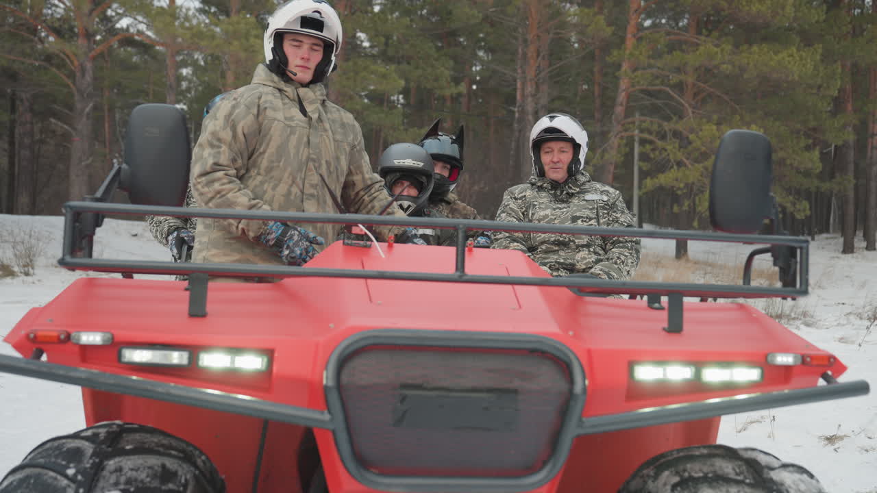 Pilota caucasico in piedi su un ATV rosso con i fari accesi, il pilota ispeziona il sentiero innevato tra i pini, due passeggeri con casco seduti in mimetica, postura concentrata e rombo del motore segnalano la prontezza per l'estremo