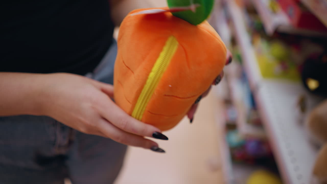 밝게 조명 된 쇼핑몰에서 녹색 세부 사항으로 활기찬 오렌지색 플러시 지갑을 들고 조사하는 여성의 클로즈업, 그녀는 다양한 물건으로 가득 찬 상점 선반 근처에 서서 부드러운 직물을 검사합니다.