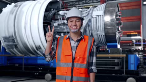asian male engineer with safety helmet smiling and showing peace gesture while standing with airplane engine maintenance conducted, analytics checking the turbine