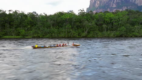 A group of people travel by longboat on a wide river through a dense jungle with a large flat-topped mountain in the background.
