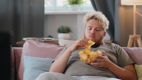 overweight man watching tv and eating chips at home