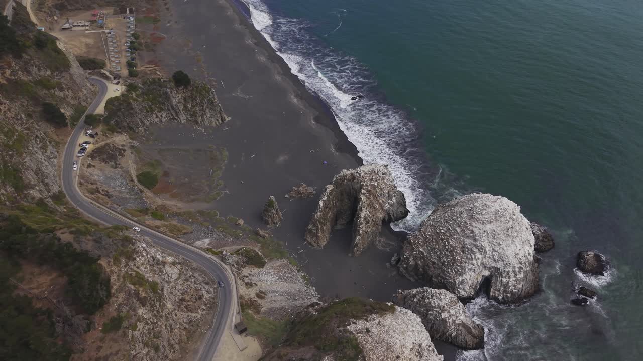 Scendendo verso le formazioni rocciose di Calabocillos lungo la costa, mostrando la strada sulla scogliera, l'area di parcheggio e una cala di sabbia nera vicino a Constitución Maule, Cile, 4K.