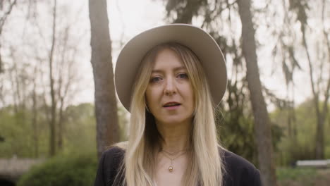 retrato de una mujer rubia con sombrero de pie en el parque y mirando a la cámara