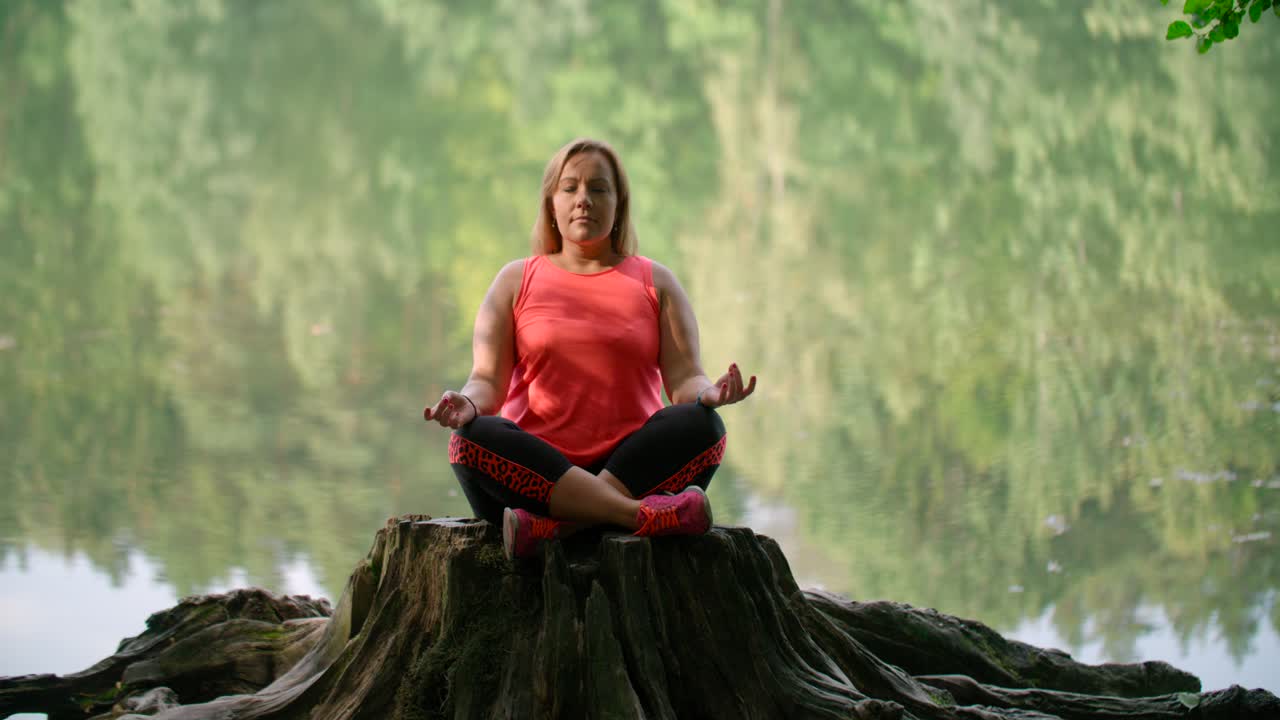 una mujer con ropa deportiva practicando yoga respiratorio en un tronco sobre el lago del bosque