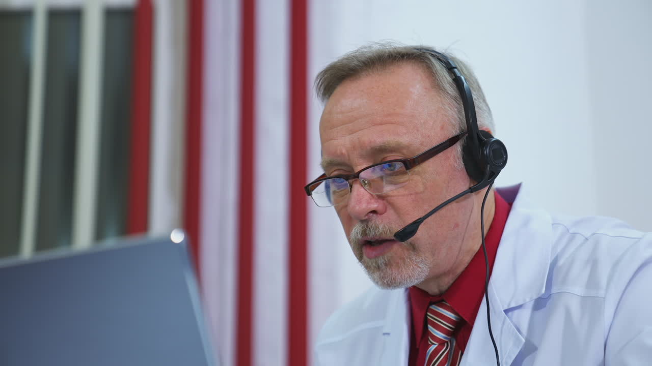 Senior physician talks to camera consulting patient online in web chat. Old adult male doctor wears white medical coat and headset makes distant video call. Telemedicine concept.