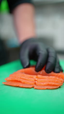 vertical footage professional culinary expert wearing black gloves precisely cutting fresh salmon fillet into delicate sashimi slices on green kitchen board for high end seafood dish