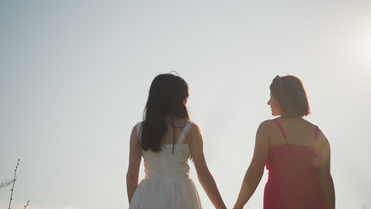 Sunlit women walk, Golden hour scene of two women walking, Backlit women with lens flare during evening walk, Silhouetted women holding hands under dramatic sky during evening meadow stroll