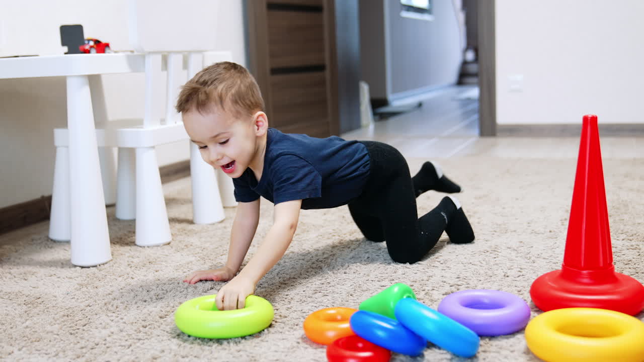 Cute baby boy plays on the carpet in his room. Cheerful kid plays with the parts of toy pyramid.