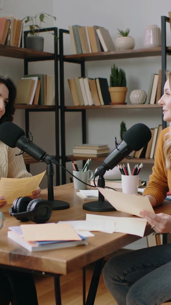 Video verticale: Conduttori in maglione leggono appunti, chinati verso i microfoni, registrando un podcast in uno studio casalingo.