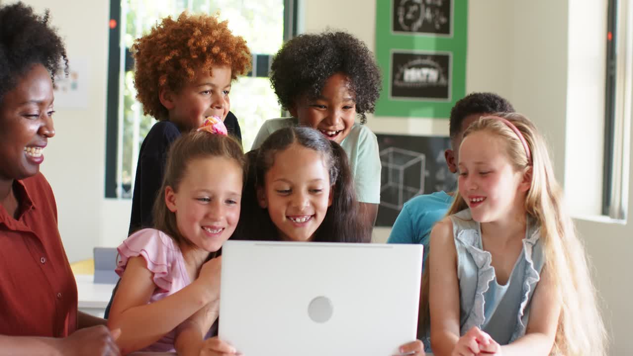 Maestra con sei bambini che reagiscono a una sorpresa su un laptop bianco, ridendo e indicando in classe.