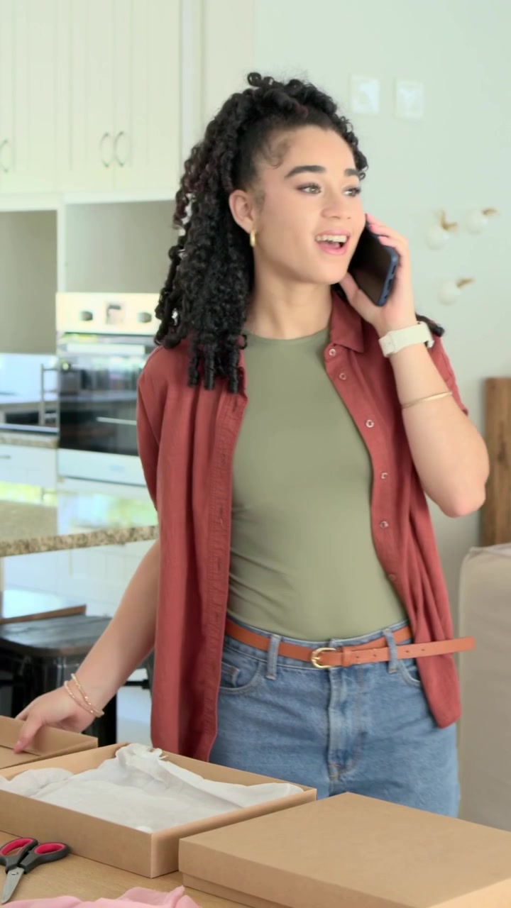 Video verticale: Una donna con maglietta color oliva risponde a una telefonata, apre scatole in cucina e prepara ordini.