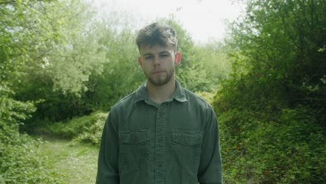 man stands in vibrant green forest, surrounded by trees in midday light, observing quietly, front portrait