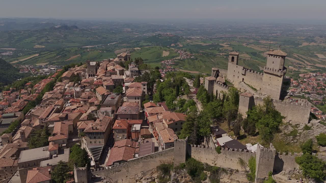 Stunning wide drone shot over San Marino showing the iconic Guaita Tower (La Rocca), historic old town streets, and Cesta Tower on Monte Titano. Clear weather and sweeping views of the countryside.