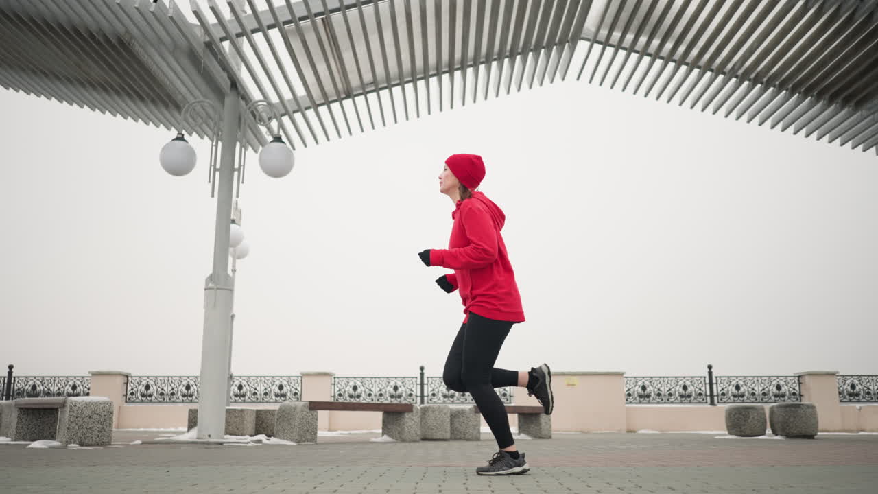 Woman in red hoodie and beanie jogging outdoors on interlocked pavement under modern pergola structure during winter, urban environment with decorative railings, benches, and snowy surroundings