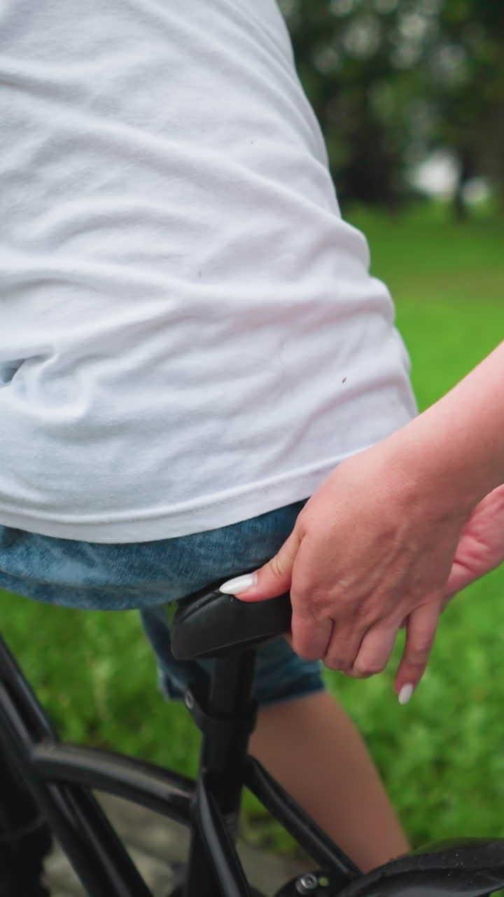 una mujer sostiene la espalda del asiento de la bicicleta de su hijo, guiándolo y apoyándolo mientras cabalga a lo largo de una pasarela cerca de un campo verde