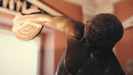 cerca de una antigua estatua masculina antigua que sostiene un disco en el brazo en un museo con luces cálidas y cambiantes con otra reliquia en el fondo borroso