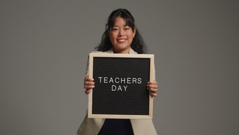 studio portrait of female teacher standing against grey background holding out notice board reading teachers day