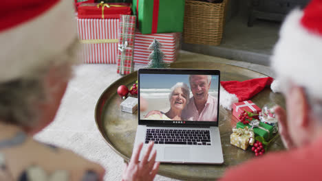 Pareja-Caucásica-Mayor-Usando-Una-Computadora-Portátil-Para-Una-Videollamada-Navideña-Con-Una-Pareja-Feliz-En-La-Pantalla