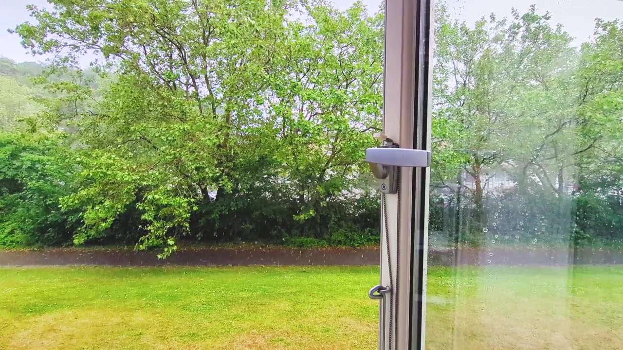 High-quality 4K footage looking through an open window during a steady rain shower in Hillerød, Denmark. Showcases lush green trees hiding a supermarket parking lot in a quiet suburban setting