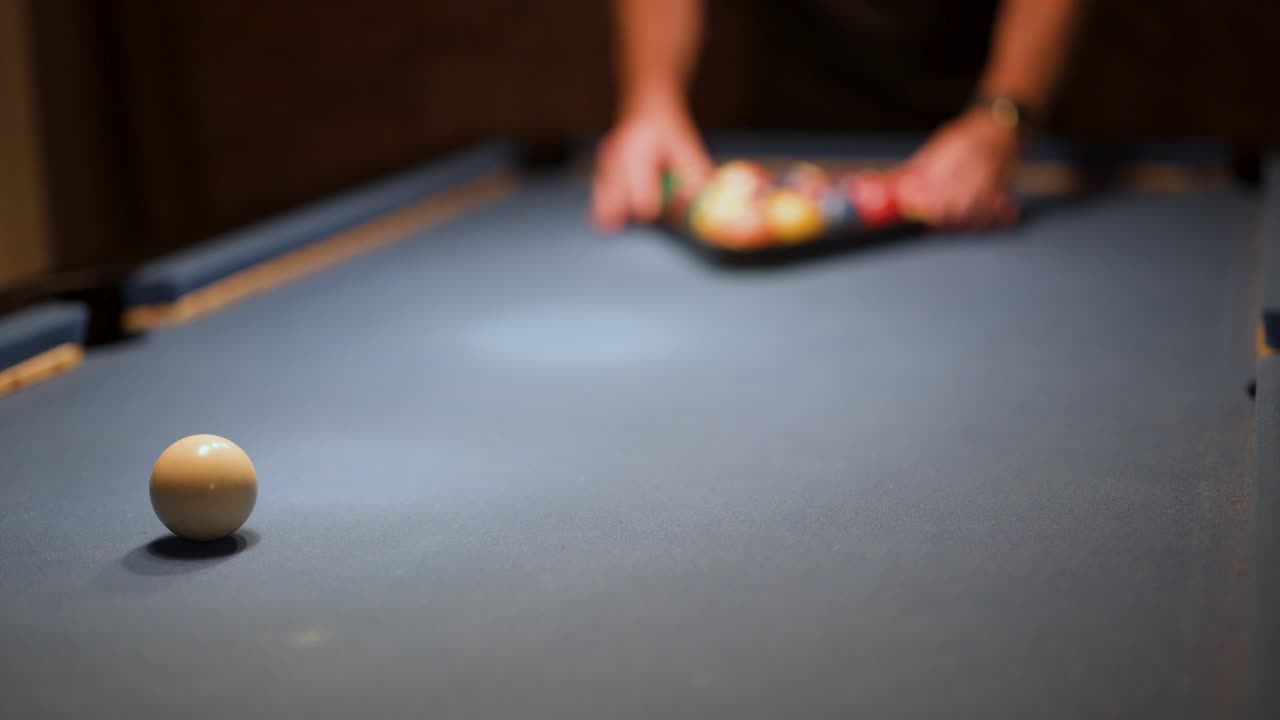 Person arranges pool balls on billiard table, low lighting, shallow depth of field, static camera