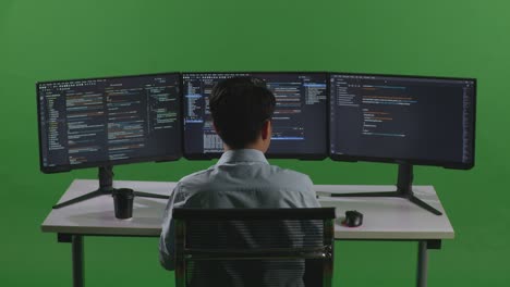 back view of asian man developer drinking coffee and using computer to write code with multiple screens in green screen studio