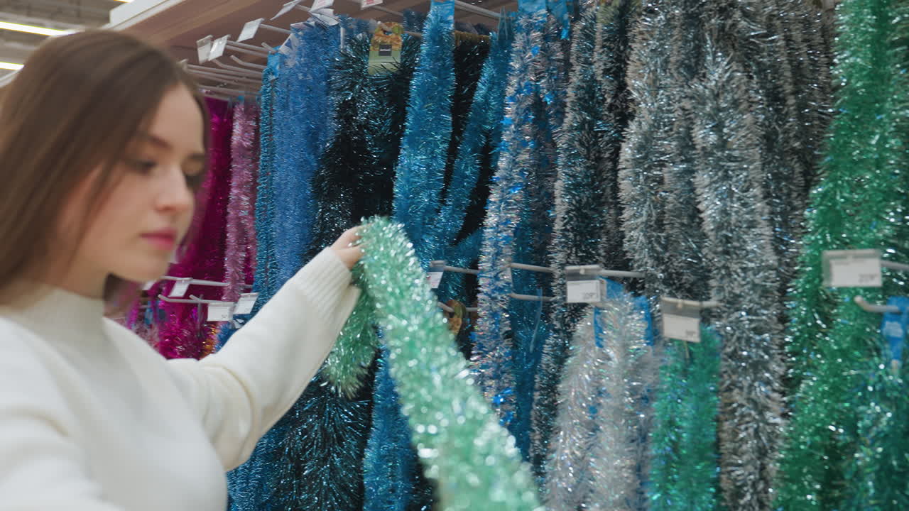 Elegant lady picks shiny green Christmas decoration from shelf in store, carefully observing it, beautiful holiday decorations on display in retail shop with vibrant colors