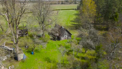 drone flying above rustic farmhouse in estonian countryside. aerial view around an old wooden cottage surrounded by forests and fields. rural village road passes around the house. beautiful spring day