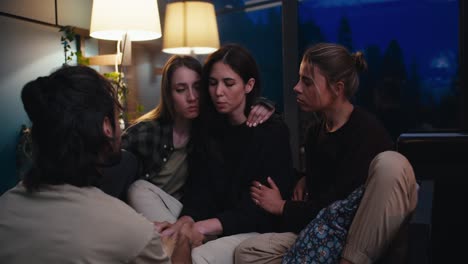 Three-girls-and-a-guy-communicate-in-the-evening-in-a-cozy-room.-A-sad-girl-talks-about-her-experiences,-and-her-friends-support-her