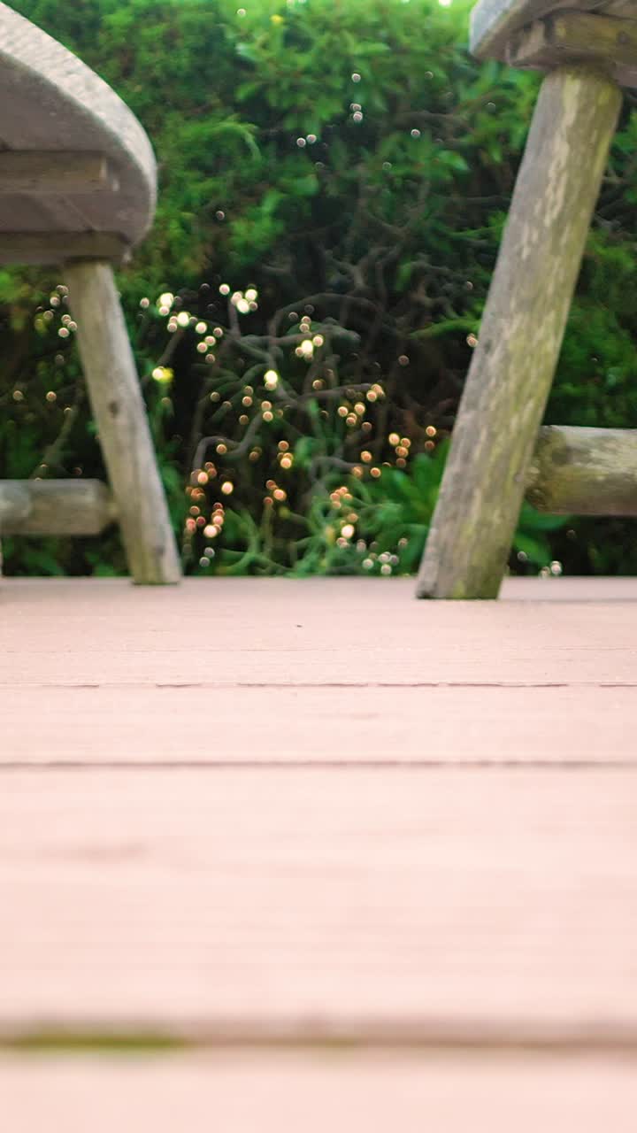 Vertical slow pan across a wooden deck with rustic outdoor chairs and soft greenery behind. Warm light and shallow depth create a cozy lifestyle scene framed for portrait video b-roll