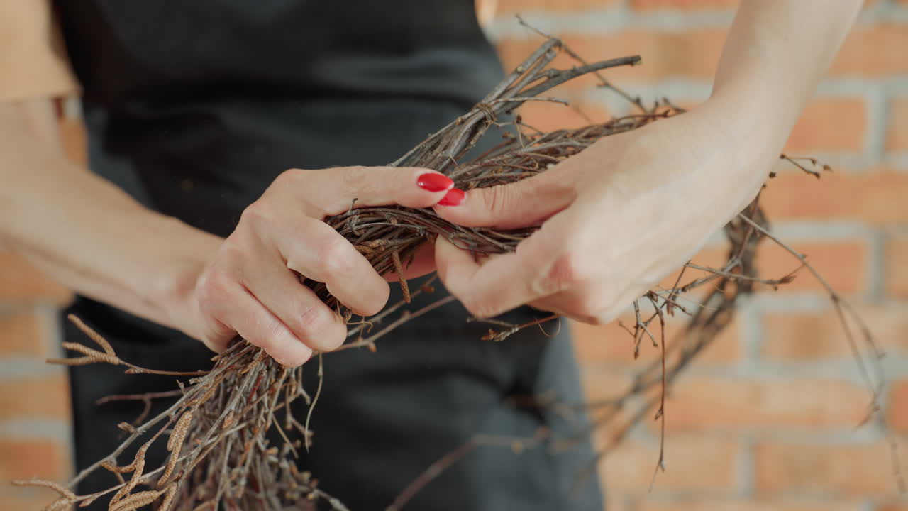 Female florist shaping dry twigs into rustic wreath decoration, hands holding bundle with red nails, creative artisan crafting natural handmade design in workshop with focus on detail and natural texture