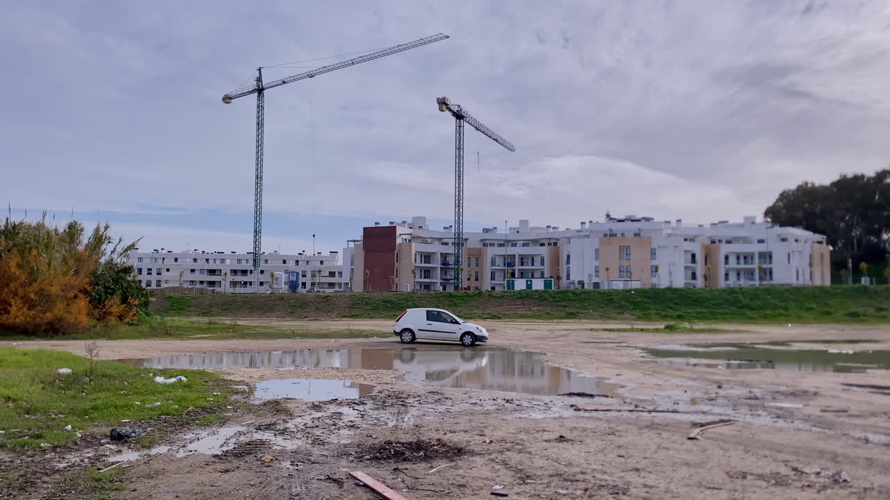 Novos edifícios residenciais em construção com gruas-torre e um carro estacionado em terreno lamacento em Sanlúcar de Barrameda, Espanha.
