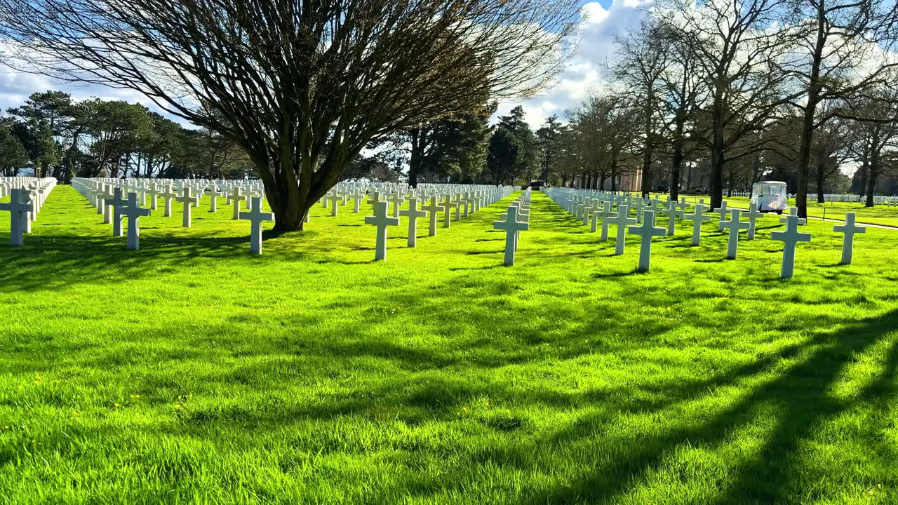 Carrellata di file infinite di croci di marmo bianco al Cimitero Americano della Normandia in Francia sotto il sole splendente con lunghe ombre degli alberi sull'erba verde lussureggiante, in onore dei soldati caduti