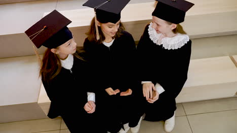 drei glückliche kleine mädchen in mütze und kleid sitzen auf der treppe und unterhalten sich bei der abschlussfeier der vorschule