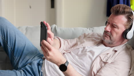 Relaxing at home, senior man using smartphone and headphones on couch