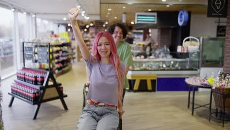 Porträt-Eines-Glücklichen-Mädchens-Mit-Rosa-Haaren,-Das-Von-Ihrem-Freund-Auf-Einem-Einkaufswagen-Geschoben-Wird,-Während-Es-Im-Supermarkt-Herumalbert-Und-Spaß-Hat