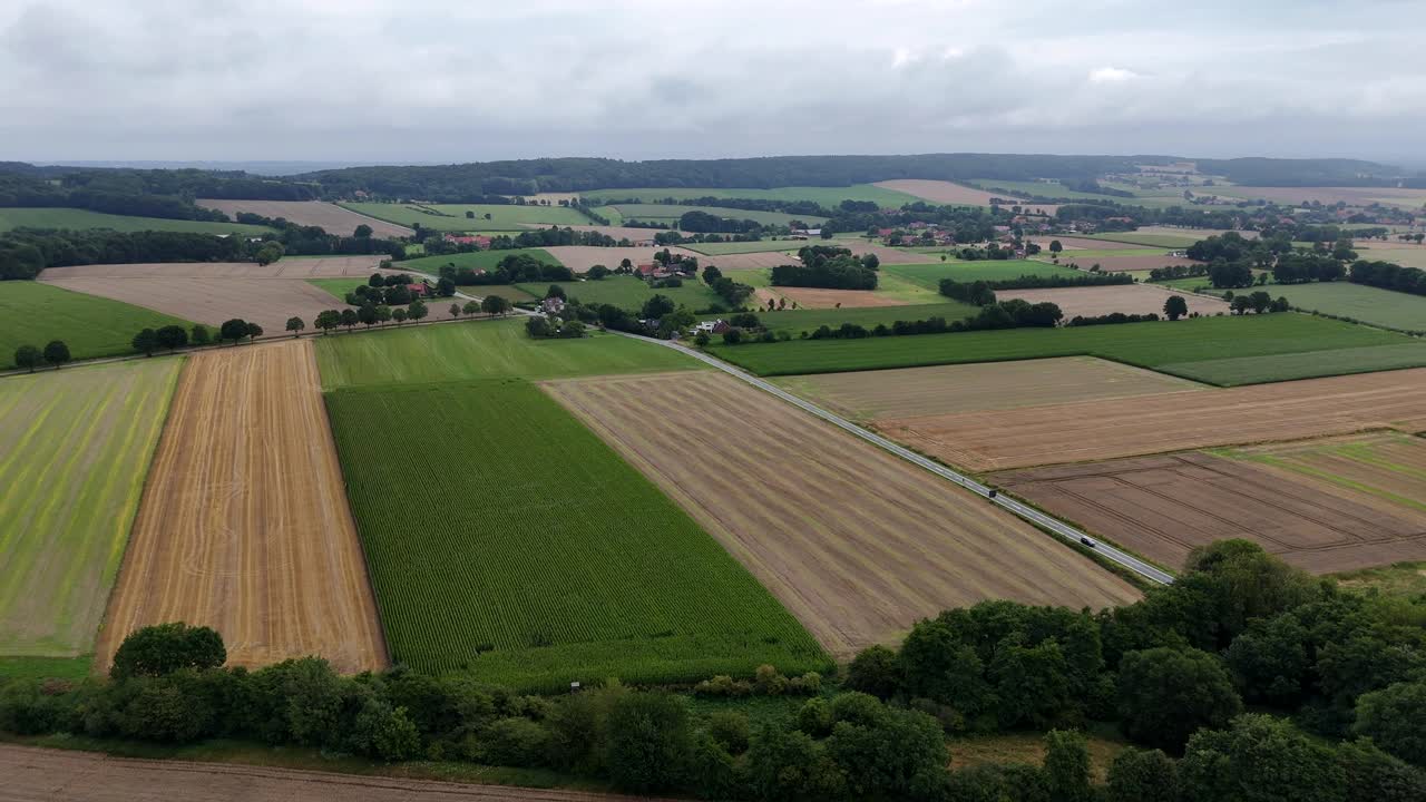 Aerial view of rural farmland in American Midwest. Patchwork fields, country roads and scattered farmhouses under a cloudy sky. Summer day with cars on rural intersection road. Wide shot